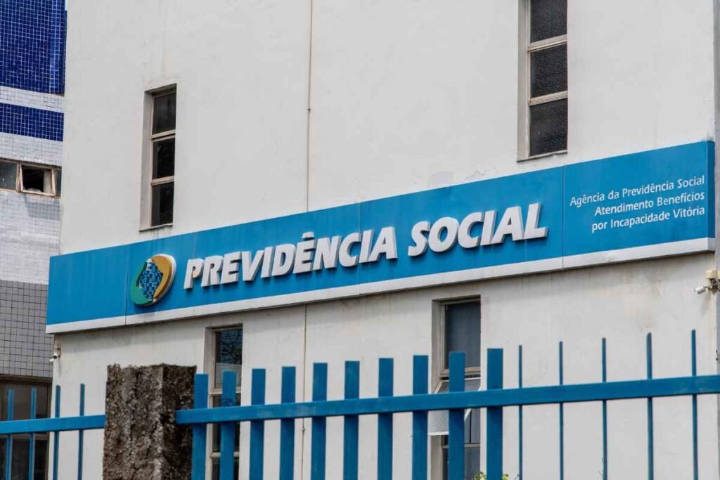 Fachada de um prédio da Previdência Social com painel azul exibindo o logotipo institucional e a inscrição “PREVIDÊNCIA SOCIAL”. À direita, há o texto “Agência da Previdência Social Atendimento Benefícios por Incapacidade Vitória”. Uma grade metálica azul ocupa o primeiro plano, e janelas estreitas são visíveis na parede branca do edifício.