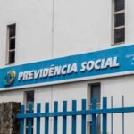 Fachada de um prédio da Previdência Social com painel azul exibindo o logotipo institucional e a inscrição “PREVIDÊNCIA SOCIAL”. À direita, há o texto “Agência da Previdência Social Atendimento Benefícios por Incapacidade Vitória”. Uma grade metálica azul ocupa o primeiro plano, e janelas estreitas são visíveis na parede branca do edifício.