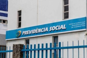 Fachada de um prédio da Previdência Social com painel azul exibindo o logotipo institucional e a inscrição “PREVIDÊNCIA SOCIAL”. À direita, há o texto “Agência da Previdência Social Atendimento Benefícios por Incapacidade Vitória”. Uma grade metálica azul ocupa o primeiro plano, e janelas estreitas são visíveis na parede branca do edifício.